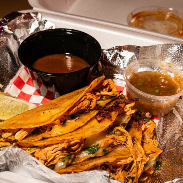 Birria tacos with lime wedges and two containers of salsa in a foil-lined takeout container.